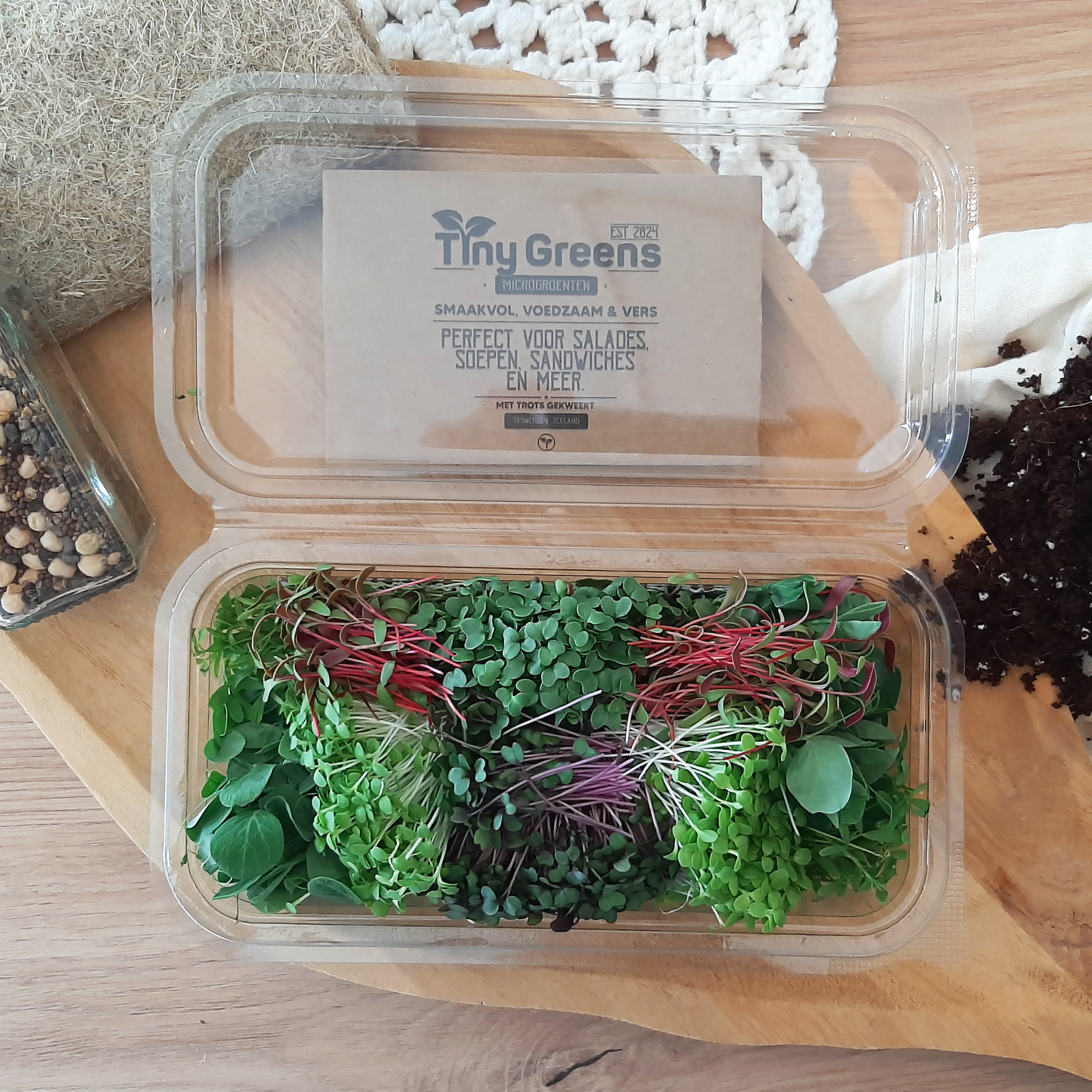 Vers geoogste microgroenten van TinyGreens, waaronder cress van snijbiet, doperwt, linzen, broccoli, rode kool en romaine bindsla. Biologisch verantwoord geteeld en rijk aan voedingsstoffen, perfect voor salades en als garnering. Gepresenteerd als bovenaanzicht in doorzichtig plastic schaal, met het label van TinyGreens er zichtbaar achter.