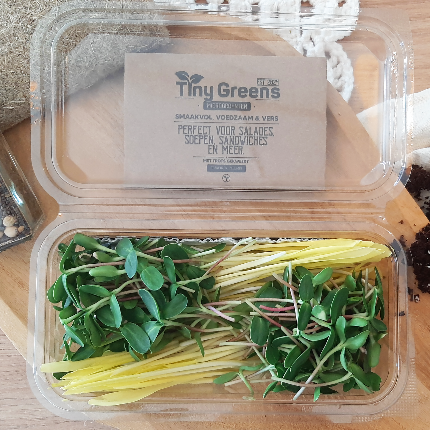 Vers geoogste microgroenten van TinyGreens, waaronder cress van zonnebloem en suikermais. Biologisch verantwoord geteeld en rijk aan voedingsstoffen, perfect voor salades en als garnering. Gepresenteerd als bovenaanzicht in doorzichtig plastic schaal, met het label van TinyGreens er zichtbaar achter.